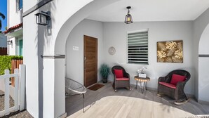Un'area esterna riparata con un muro bianco, una porta in legno, sedie in vimini con cuscini rossi, un tavolino e una lampada a sospensione.