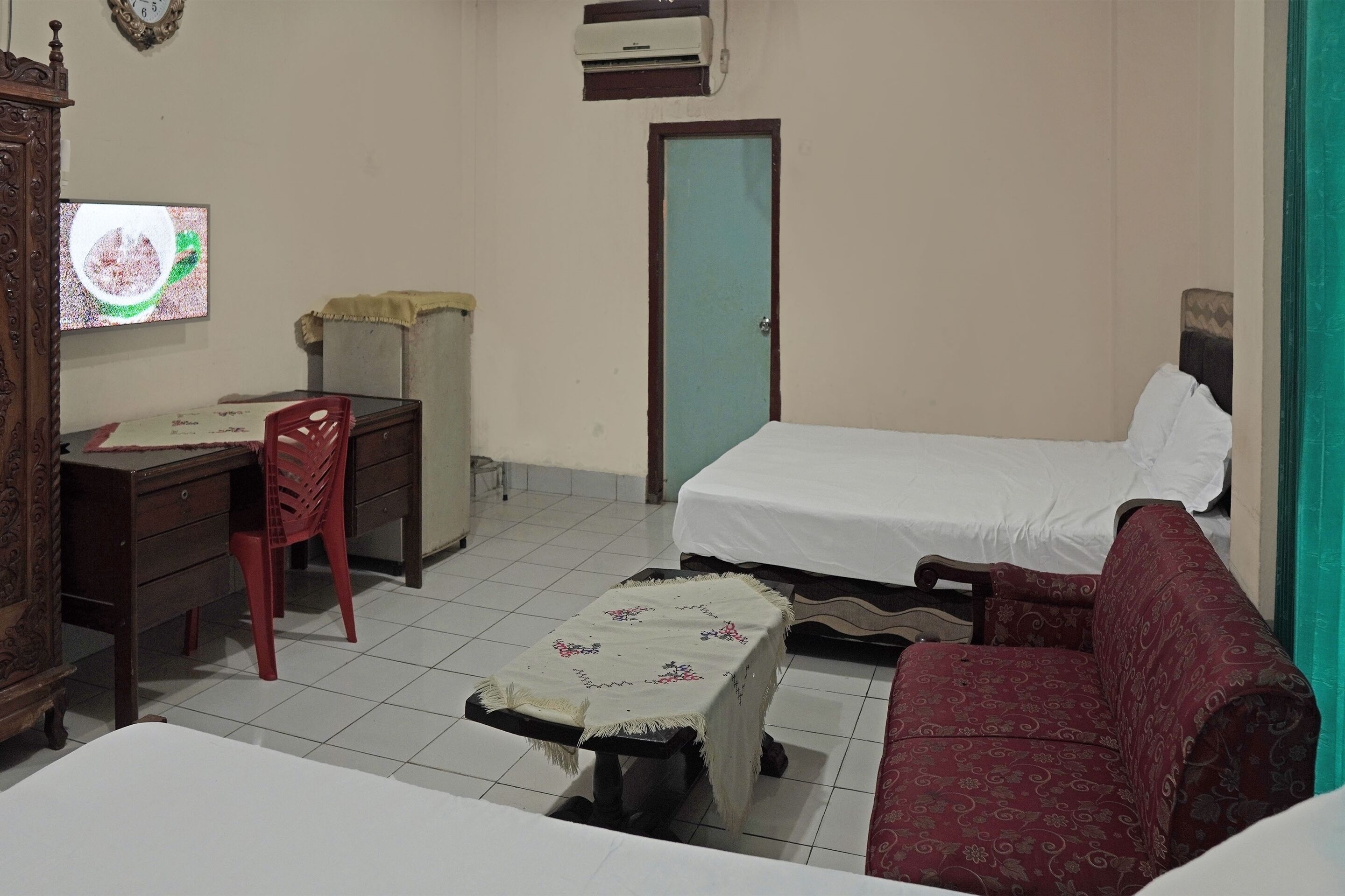 A room with a bed, a desk with a chair, a small table with a tablecloth, a red sofa, and a television on the wall.