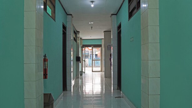 A corridor with tiled floors and walls, leading to an open area with a door and windows. - Capital O 92958 Hotel Raflessia Syariah (Lawang Kidul)