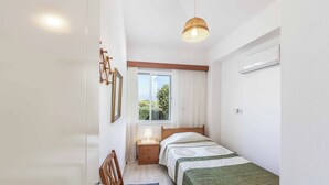 A small, well-lit bedroom with a single bed, a bedside table with a lamp, a window with curtains, and a hanging light fixture. - Villa Chara Mari (Neo Chorio)