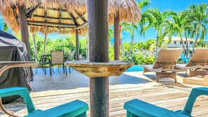 A wooden deck with lounge chairs and a grill area, surrounded by palm trees and a clear view of the ocean. - Happy Tingler With Pool Tiki and 100 ft of Dockage Right Next to Sombrero Beach (Marathon)