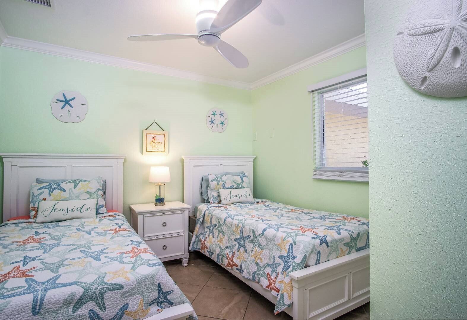 A bedroom with two beds, a ceiling fan, a nightstand with a lamp, and a window with blinds.