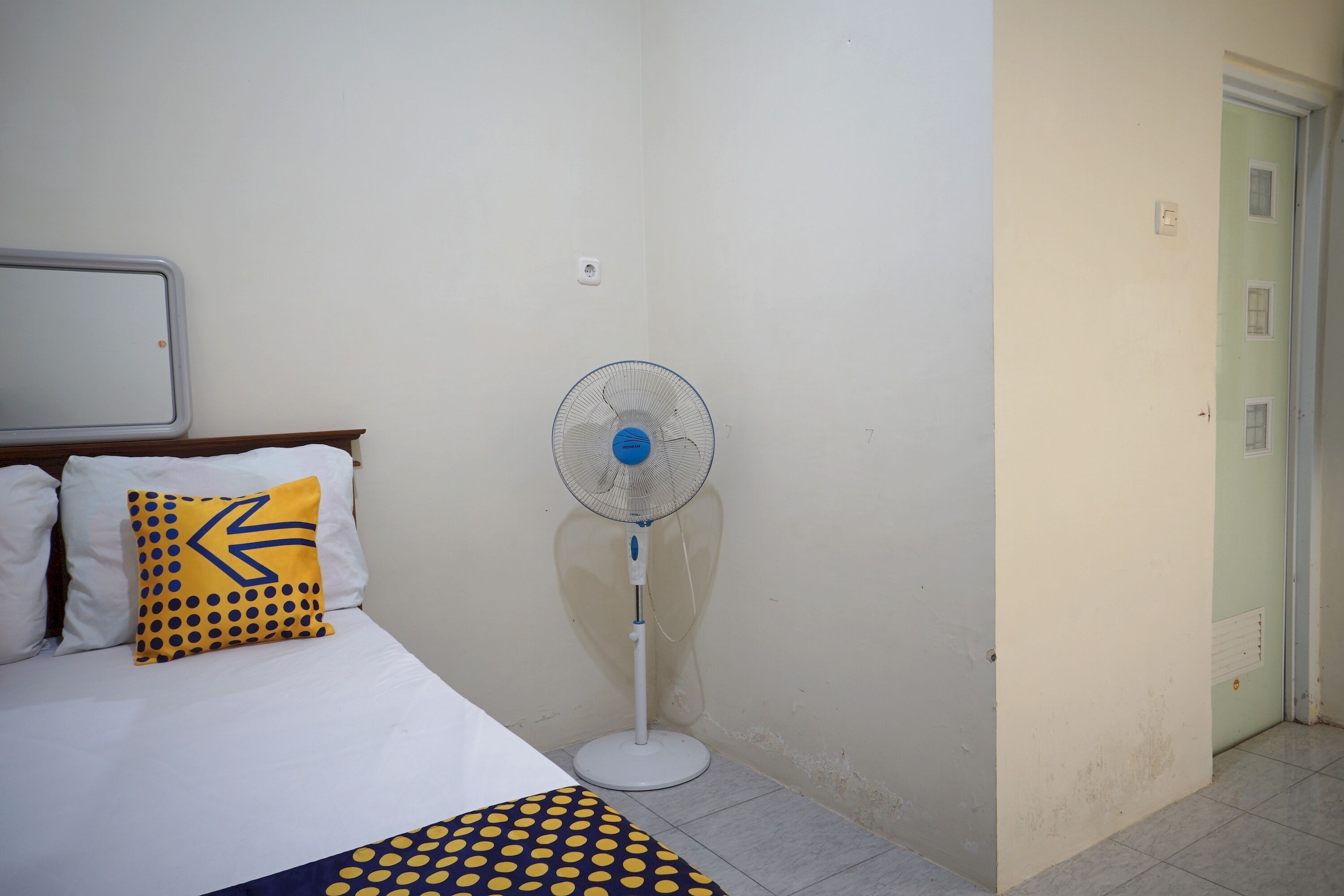 Une chambre d’hôtel avec un lit, un ventilateur sur pied et une porte.