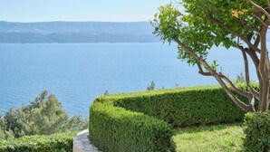 A landscaped area with a neatly trimmed hedge, a tree with green leaves, and a view of a large body of water. - Bella Vista Dalmatien #1 (Celina)