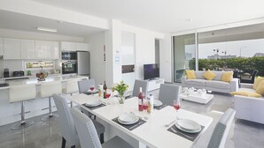 Une cuisine moderne avec des meubles blancs, un coin repas avec une table blanche dressée pour quatre personnes, et un salon avec un canapé et un téléviseur.