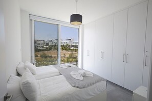 Ein modernes Schlafzimmer mit einem großen Bett, einem weißen Kleiderschrank und Blick auf Gebäude durch eine Schiebetür.