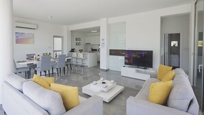 Un salon moderne avec un téléviseur à écran plat, des canapés gris agrémentés de coussins jaunes, une table basse et un coin repas avec des chaises, et une cuisine en arrière-plan.