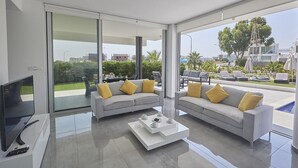 Un salon moderne doté d’un grand téléviseur, de deux canapés gris avec des coussins jaunes, d’une table basse blanche et d’une porte vitrée donnant sur un balcon aménagé avec du mobilier d’extérieur.