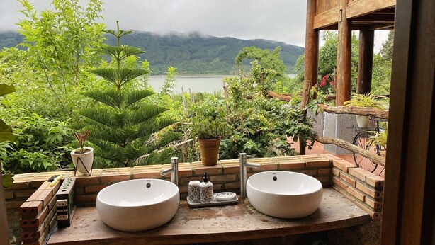 A bathroom with two white sinks, a wooden counter, and a view of a lake and mountains. - Khu nghi duong Kala Campark (Bao Thuan)