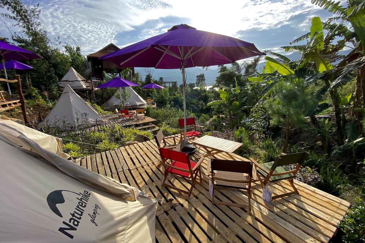 Un espacio al aire libre con temática natural, que incluye una terraza de madera, sombrillas púrpuras y tiendas de campaña blancas.