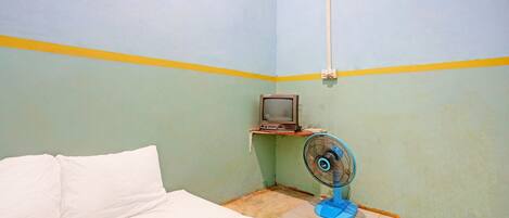 Una habitación pequeña y sencilla con una cama, un televisor en un estante y un ventilador de pie.