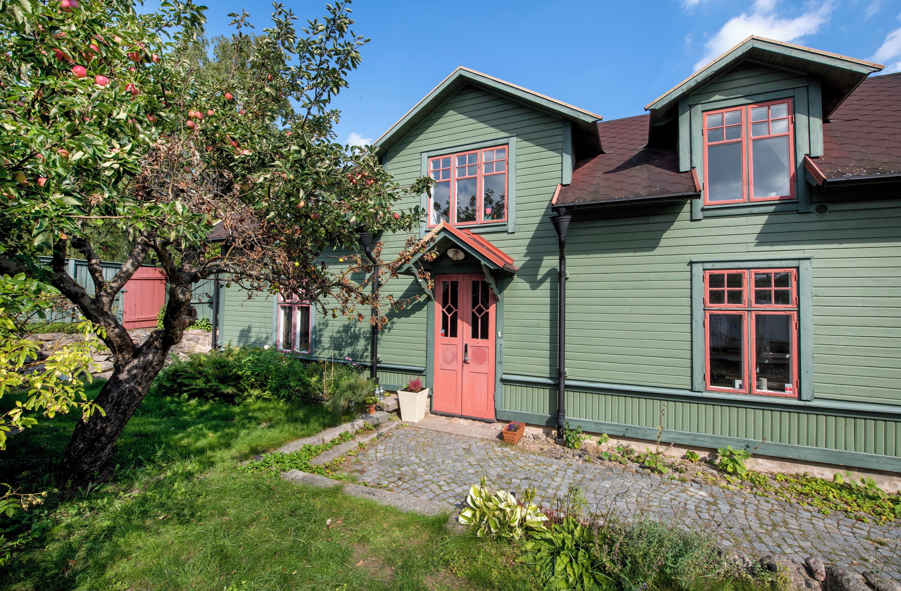 A green house with a red door, a small porch, and a tree with red apples.