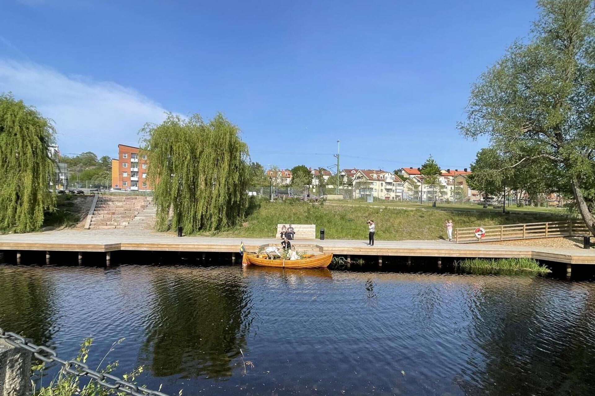 A riverside walkway with a wooden platform, a small boat, and a bridge.