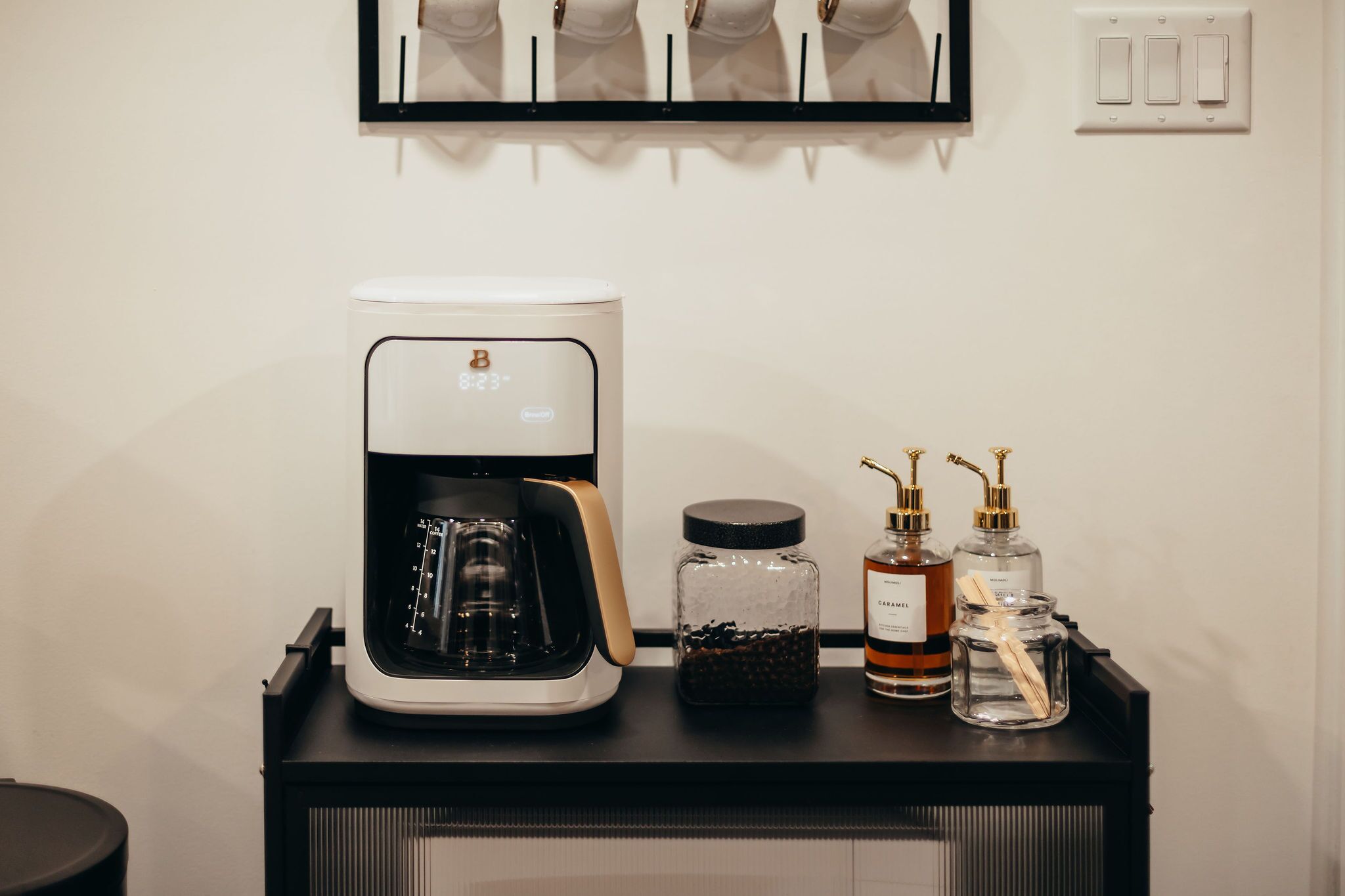 En kaffemaskine, en krukke med kaffebønner og to parfumeflasker på et sort fad.