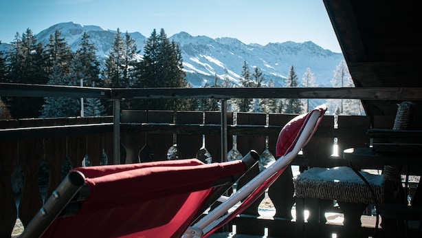 A wooden deck with red lounge chairs and a cushioned bench, overlooking snow-capped mountains and evergreen trees. - Malixerhof (Churwalden)