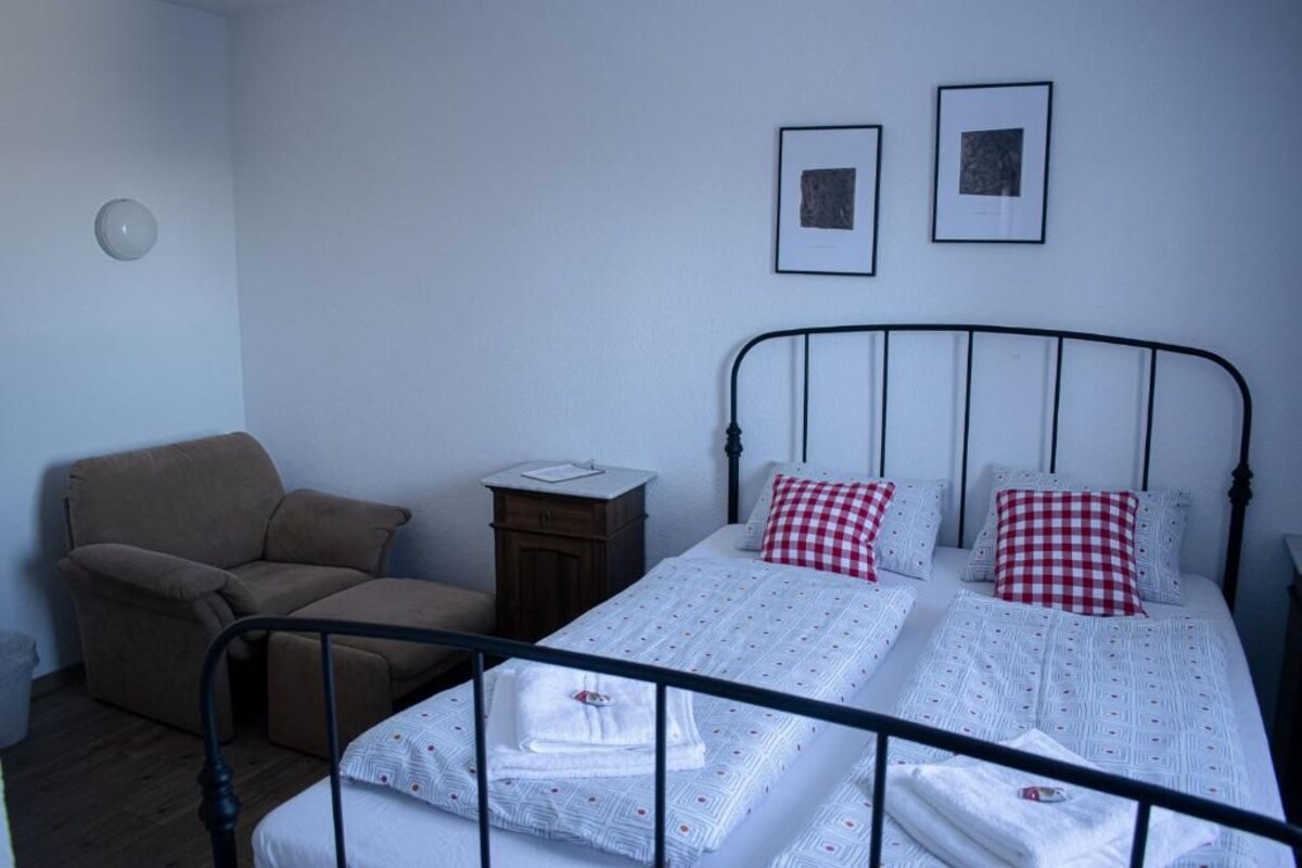 Un dormitorio con cama, una silla, una mesita de noche y dos cuadros en la pared.