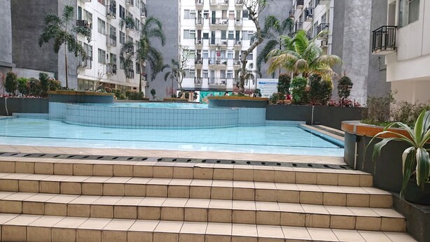 A swimming pool with tiled steps leading up to it, surrounded by palm trees and a multi-story building with balconies. - Collection O 92981 Apartemen The Jarrdin By Gold Suites PropertyNearSTEI ITB: Sekolah Teknik Elektro dan Informatika ITB (Bandung)