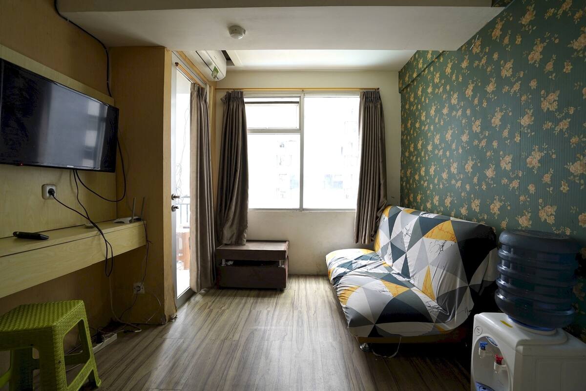 A room with a bed, a TV mounted on the wall, a desk with a computer, a water dispenser, and a window with curtains.