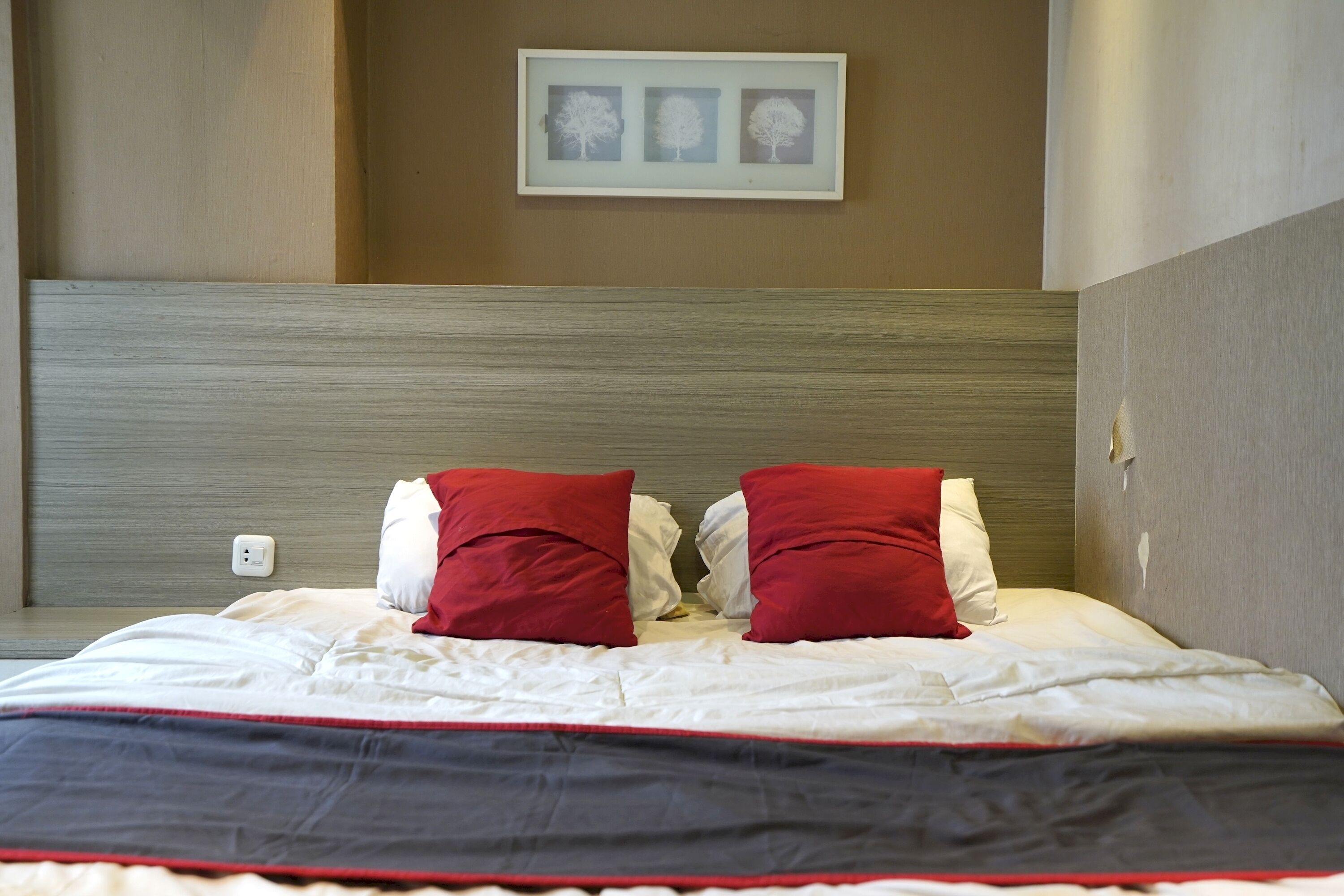 Una cama bien hecha con almohadas rojas y blancas, un cuadro enmarcado encima y un interruptor de luz en la pared.