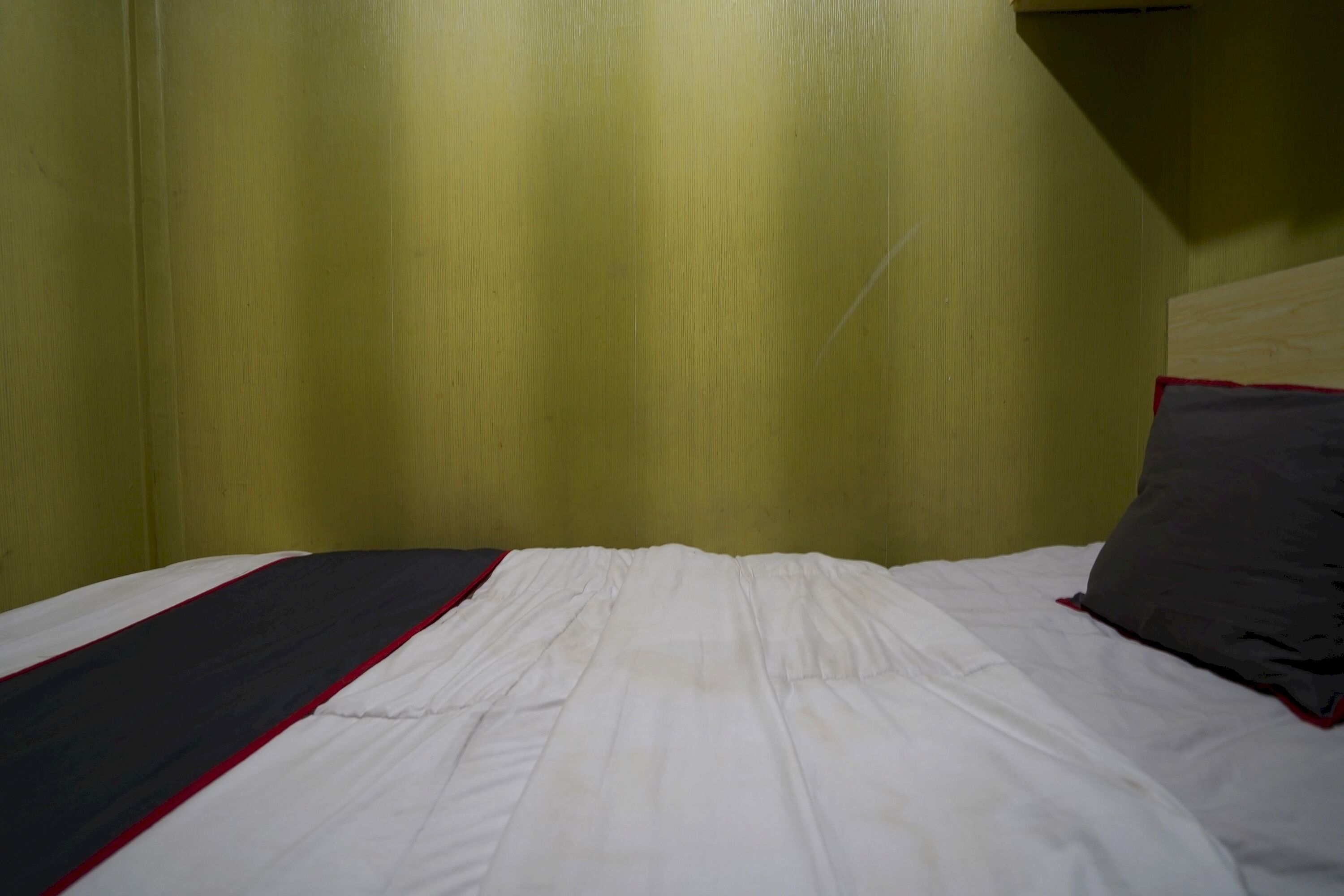 A bed with white bedding and a red stripe, a dark pillow, and a green wall.