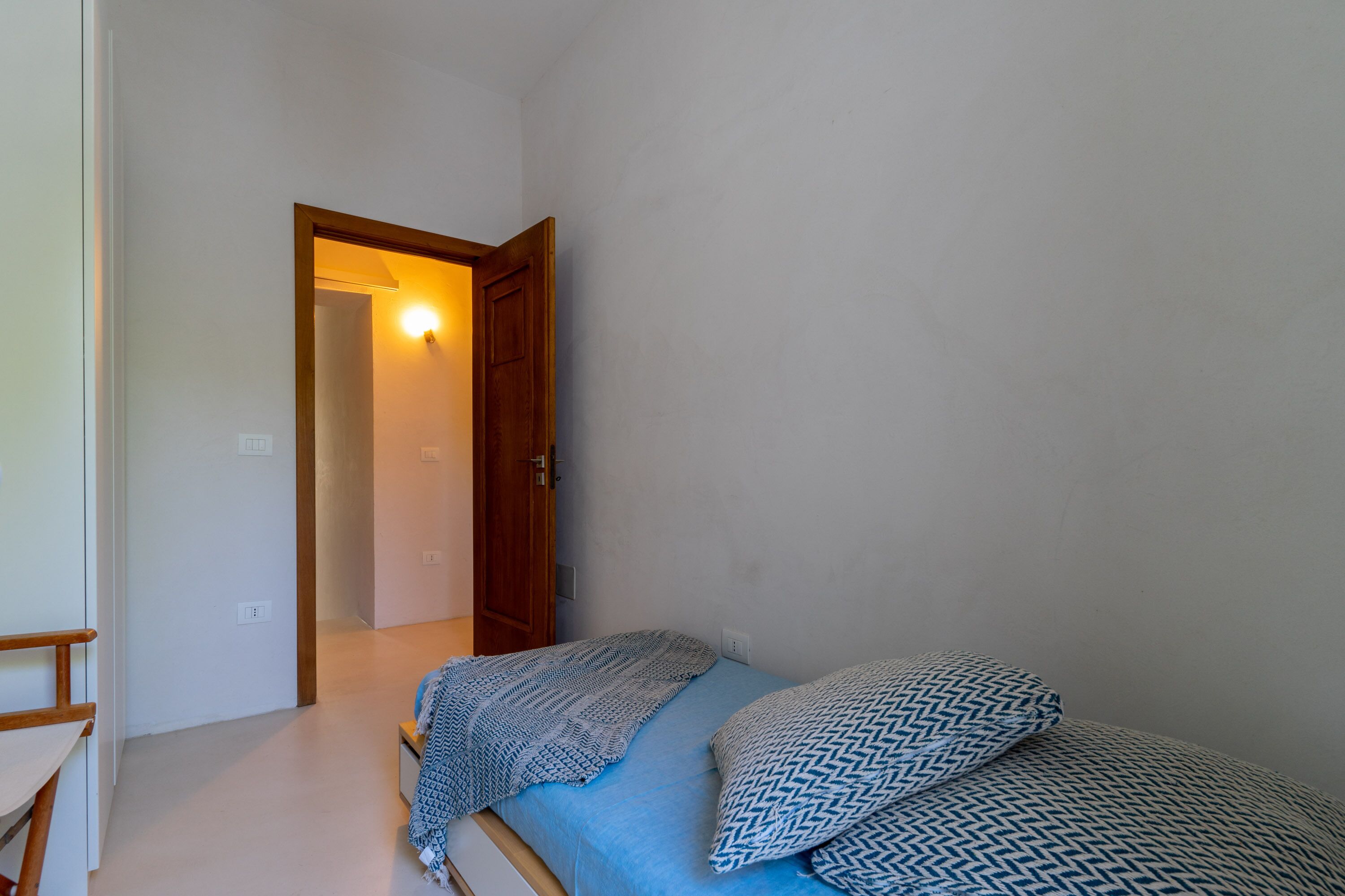 Ein kleines Zimmer mit einem Einzelbett, einer Holztür, einem Lichtschalter und einer geblümten Decke.