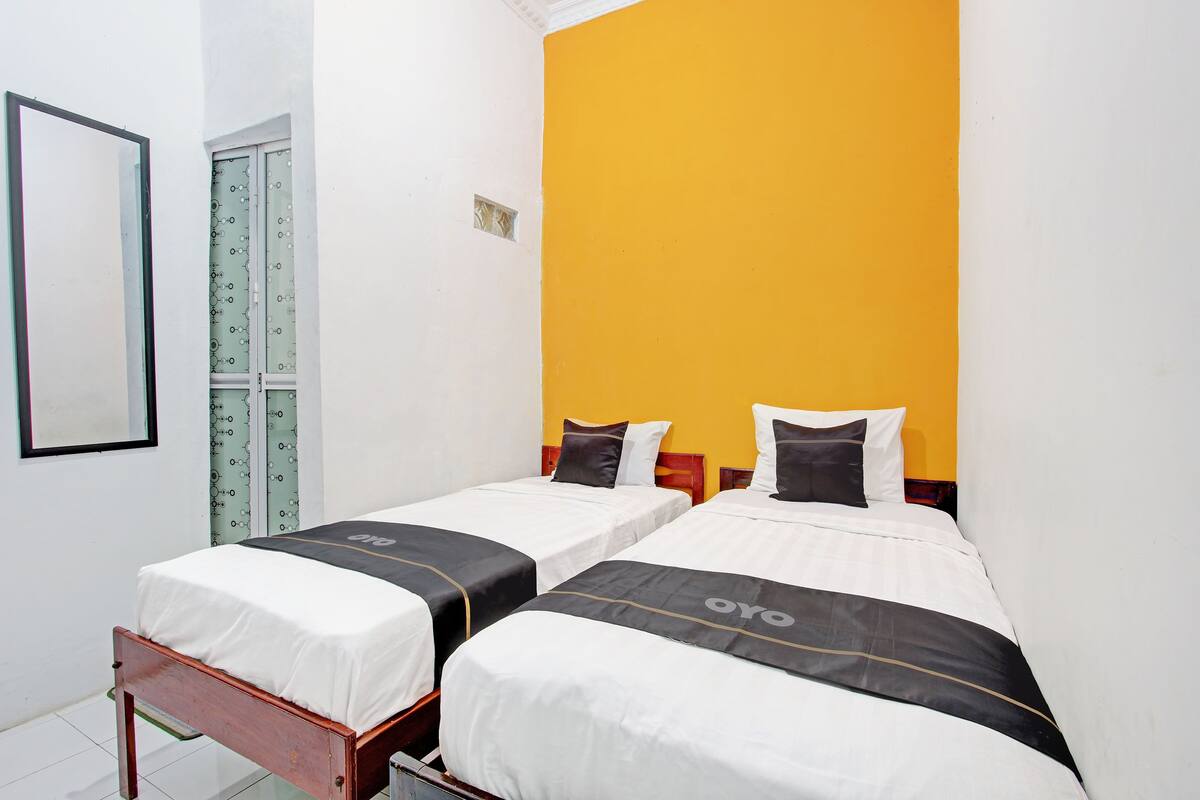 A hotel room with two single beds, a small window, and a plain white wall.