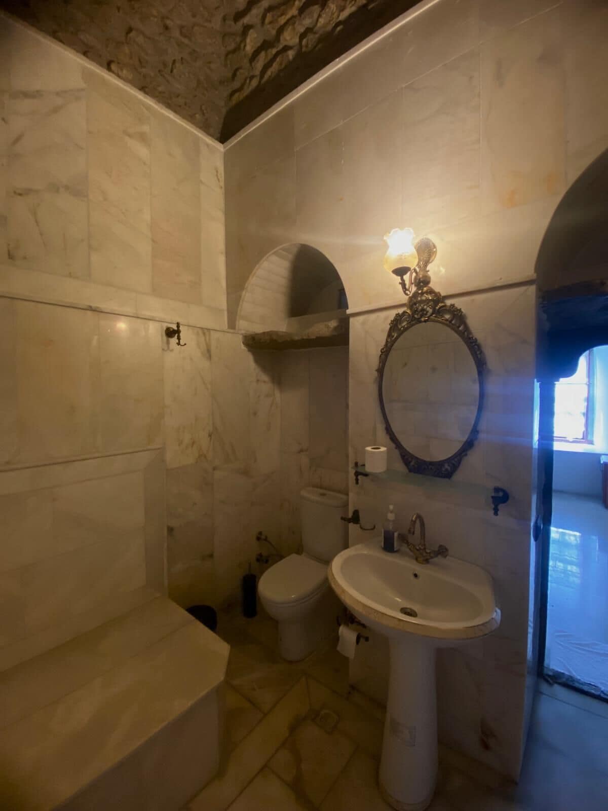 A bathroom with a marble floor, a pedestal sink, a toilet, and a mirror with an ornate frame.