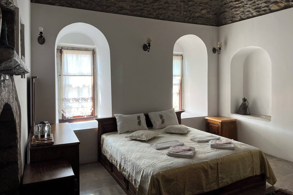 A bedroom with a bed, two windows with arched tops, a nightstand, and a lamp.