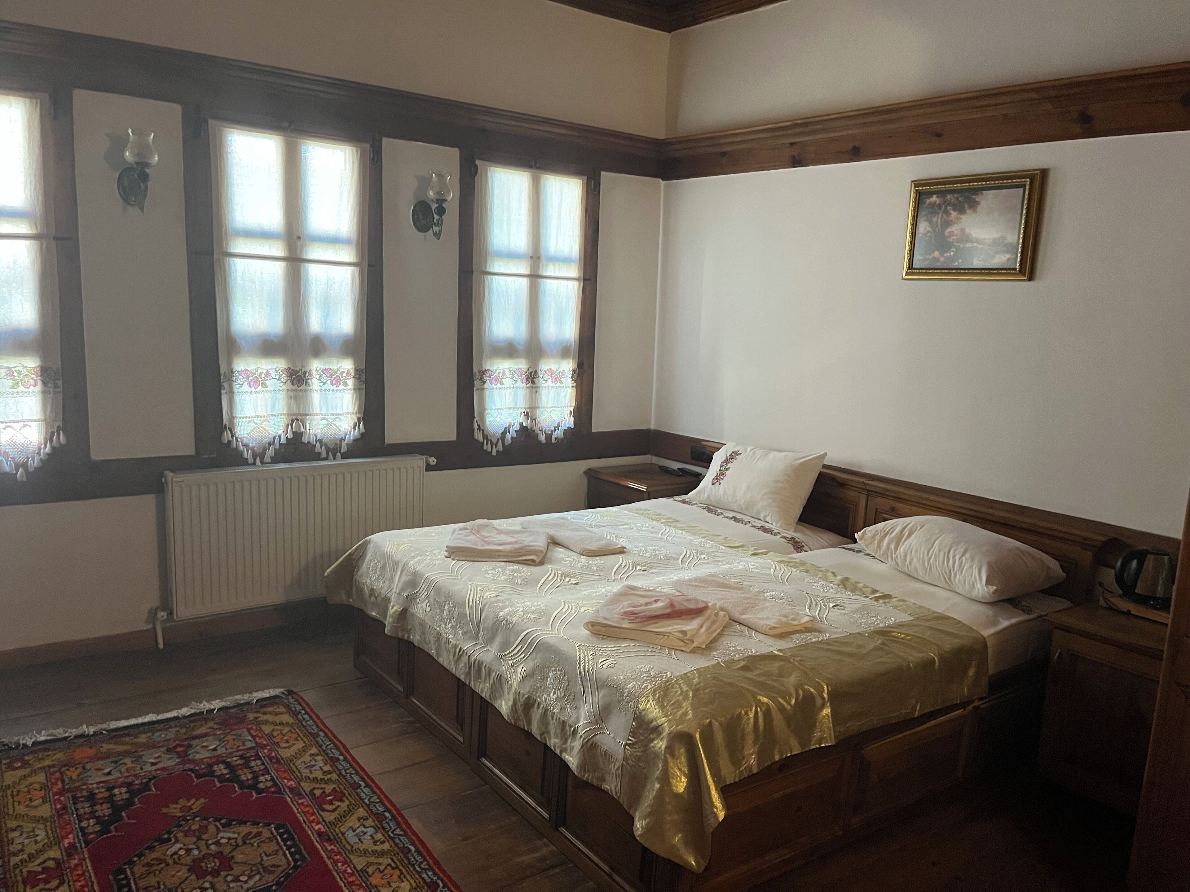 A bedroom with a wooden bed, a nightstand, a framed picture on the wall, and a window with lace curtains.
