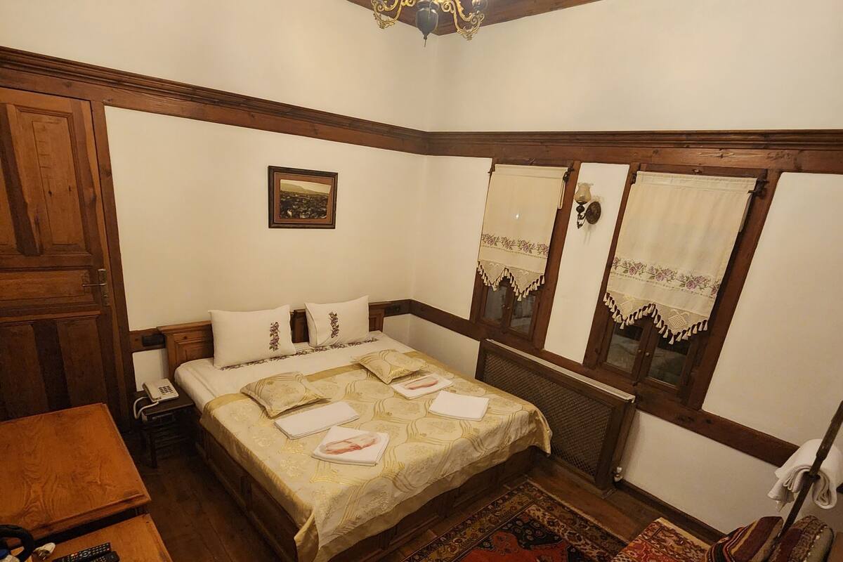 A traditional room with a wooden ceiling, a bed with a patterned bedspread, a wooden nightstand, a telephone, a chair, and a window with curtains.