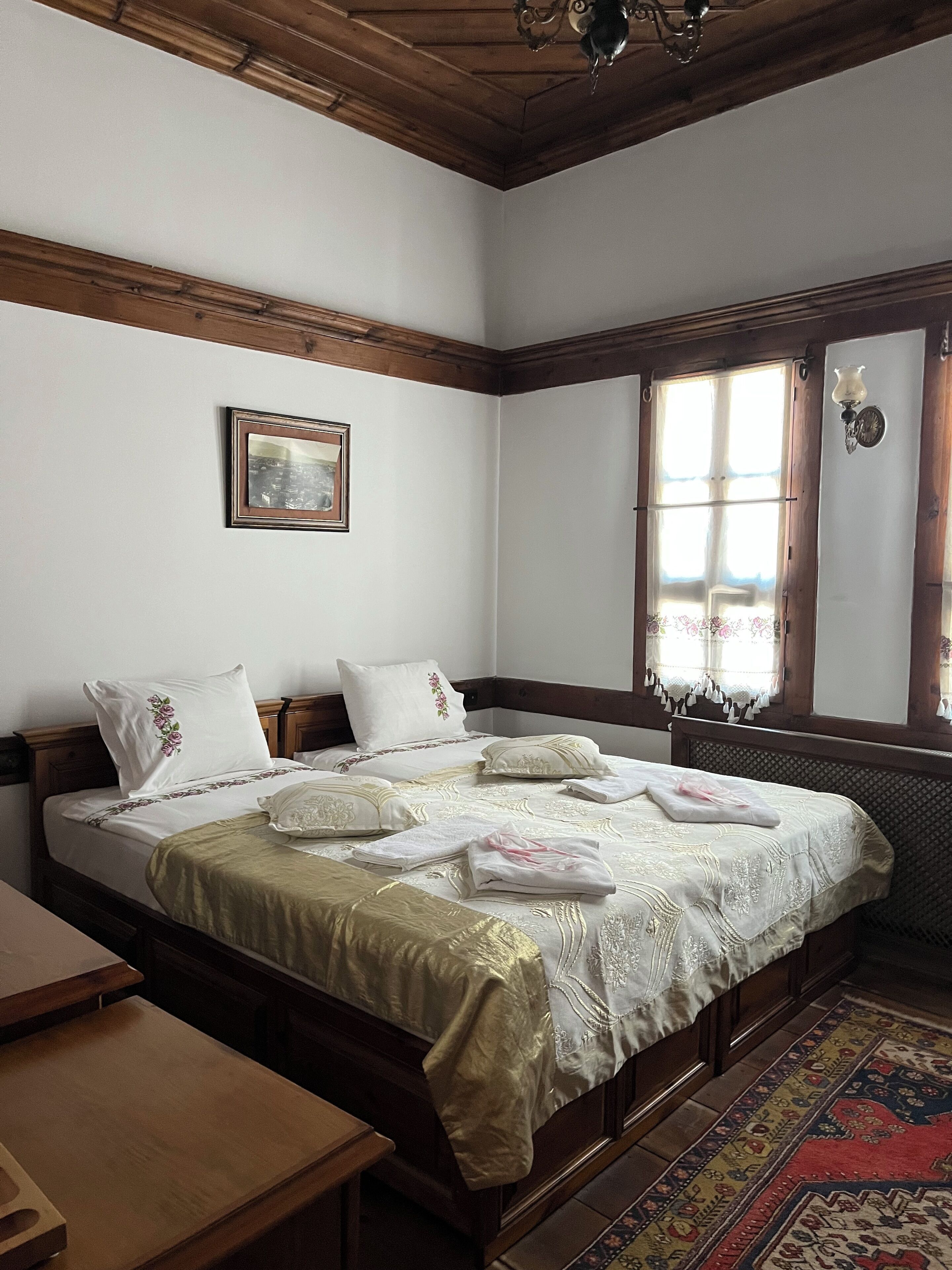 A bedroom with a wooden bed, a window with curtains, a bedside table, and a small rug.