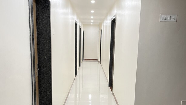 A long, narrow hallway with white walls and a shiny tiled floor. There are doors on both sides, and a set of stairs with brown steps and a handrail on the right. - Hotel Rajdhani (Latur)