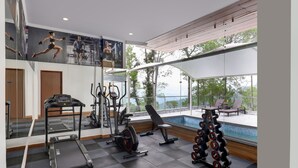 A gym with a treadmill, elliptical, bench, and dumbbells, large windows with a view of trees and a pool. - Lemon Tree Hotel Mcleodganj (Dharamshala)