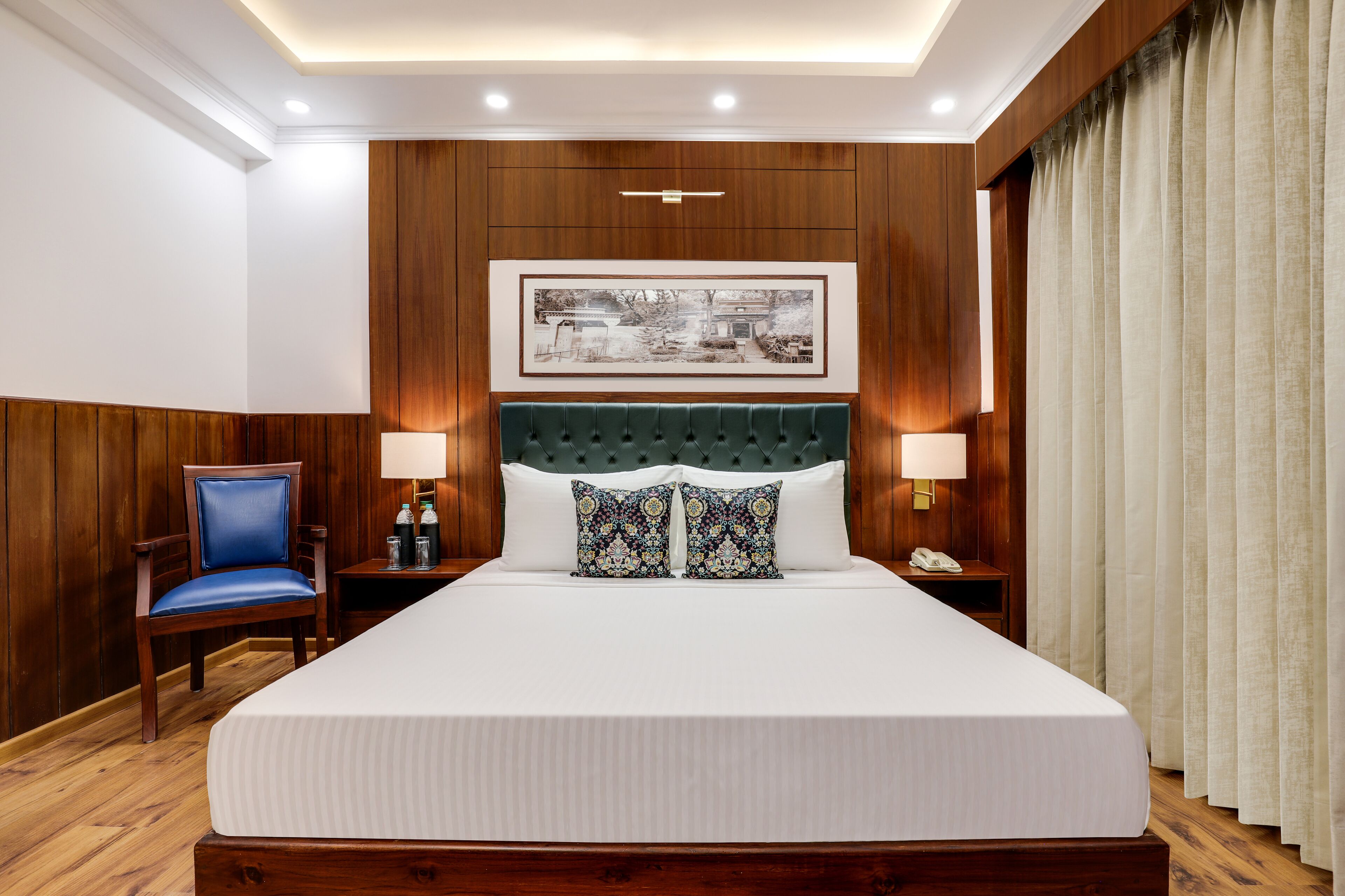 A hotel room with a large bed, wooden panel walls, a framed picture above the bed, two bedside lamps, a chair, and a nightstand with a phone.