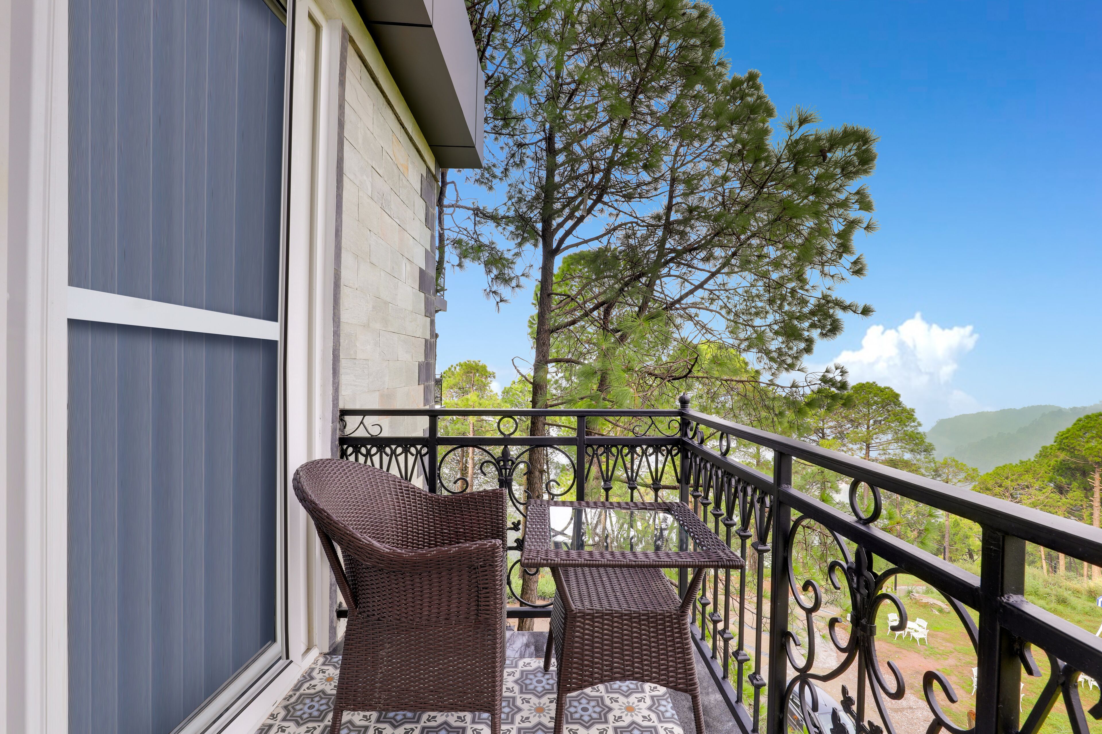 A balcony with wicker furniture and a patterned floor, overlooking a scenic landscape with trees and hills.