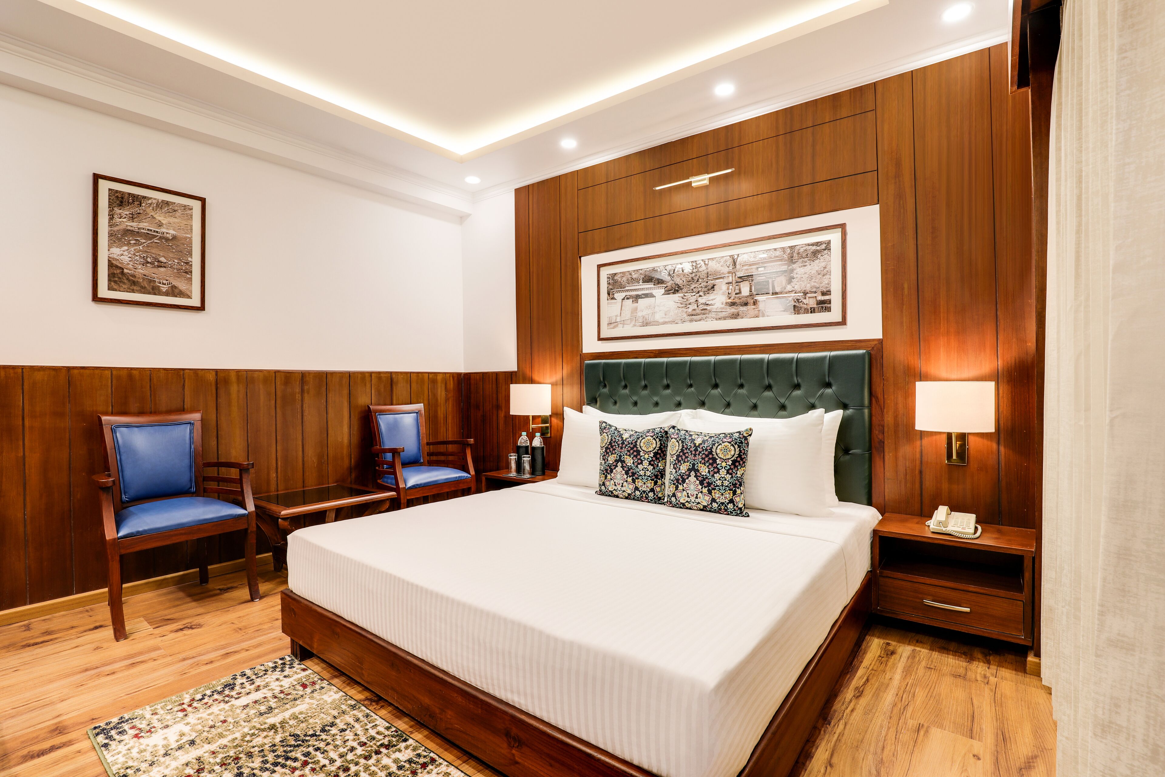A hotel room with a large bed, two nightstands, a green upholstered headboard, and wooden paneling on the walls.