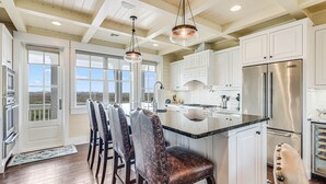 A kitchen with white cabinets, a stainless steel refrigerator, and a bar area with leather chairs. - Stunning Lakeside Home near Keira’s Pool & More Cinnamon Shore Amenities. (Port Aransas)