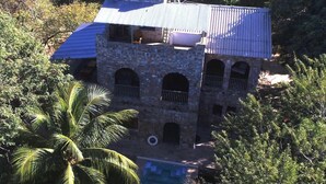 A stone building with a blue roof, surrounded by lush greenery and a clear view of the sea.