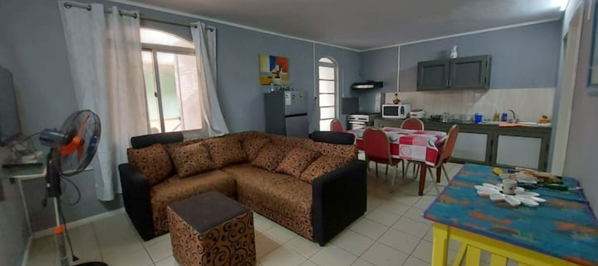A living room with a sofa, a dining table with chairs, a kitchen area with cabinets and a microwave, and a fan.