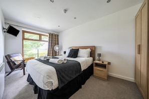 A hotel room with a bed, a chair, a bedside table, a wardrobe, a TV mounted on the wall, and a window with curtains. - Modern Peaceful Open Plan 1-bed Apartment Rutland (Oakham)