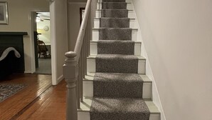 Una escalera con peldaños alfombrados y barandillas blancas, que conduce a una plataforma con una lámpara en el techo.