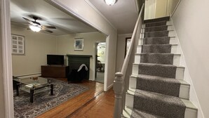 Amplia sala de estar con piso de madera, chimenea, una escalera con escalones alfombrados y un ventilador de techo.