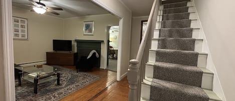 A spacious living room with hardwood flooring, a fireplace, a staircase with carpeted steps, and a ceiling fan.