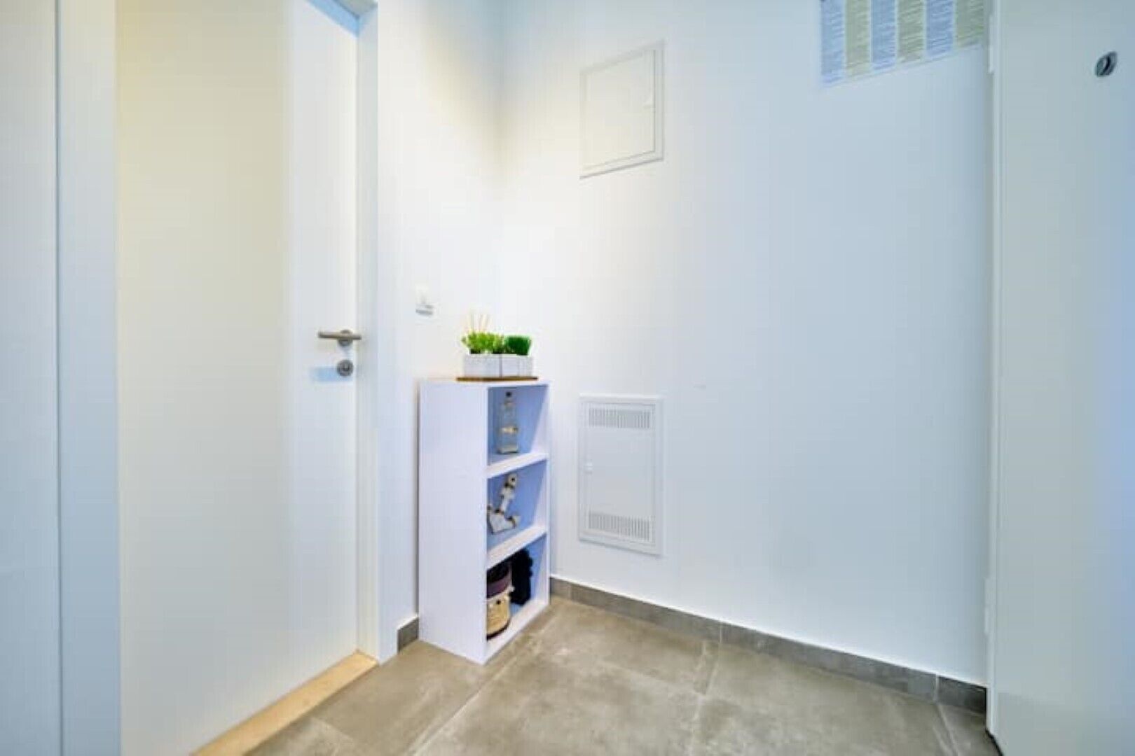 A hallway with a white door, a white shelf with a plant, and a vent on the wall.