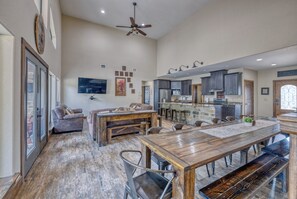 Amplia sala de estar con una mesa de comedor de madera, asientos cómodos, una cocina con armarios oscuros y un ventilador de techo.