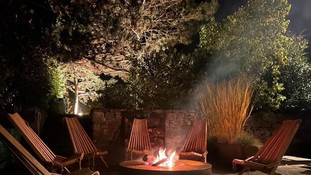 A circular outdoor fire pit with four wooden lounge chairs around it, set against a stone wall with vegetation.