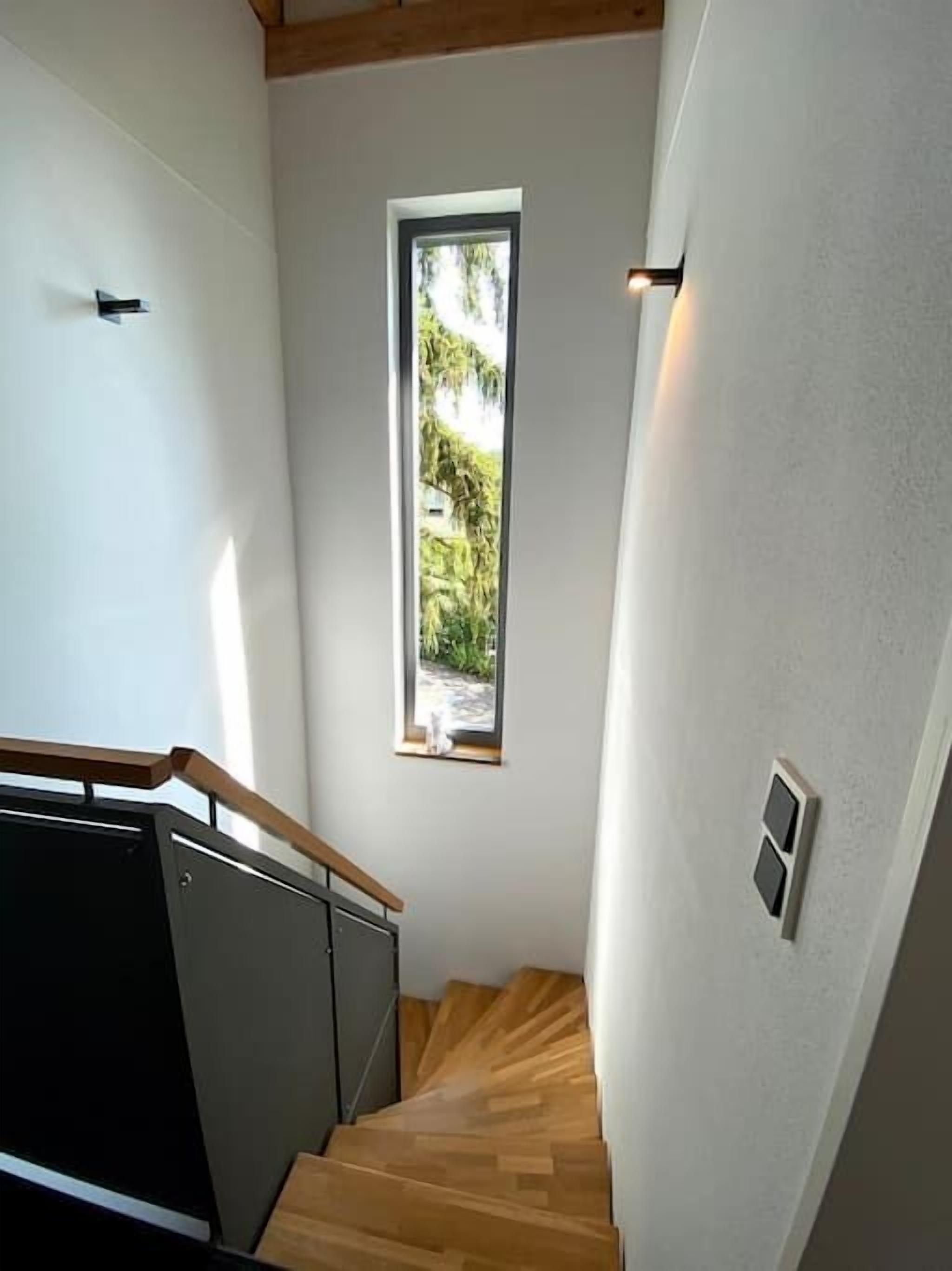 Eine Treppe mit Holzdielen und einem schwarzen Geländer, die zu einer Zwischenebene mit einem kleinen Fenster mit Blick auf Grün führt.