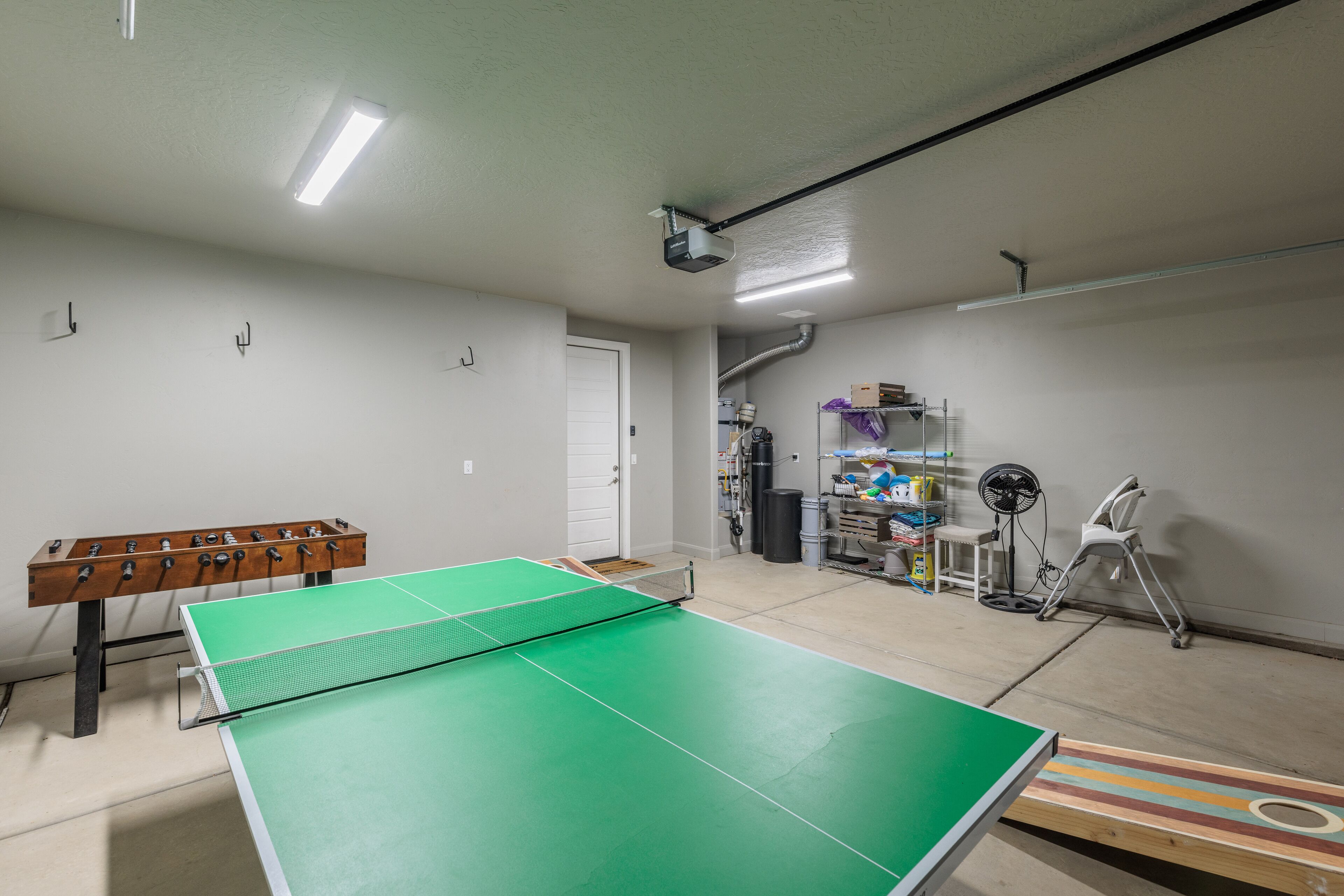 A ping pong table in a room with a white door, a shelf with various items, and a fan.