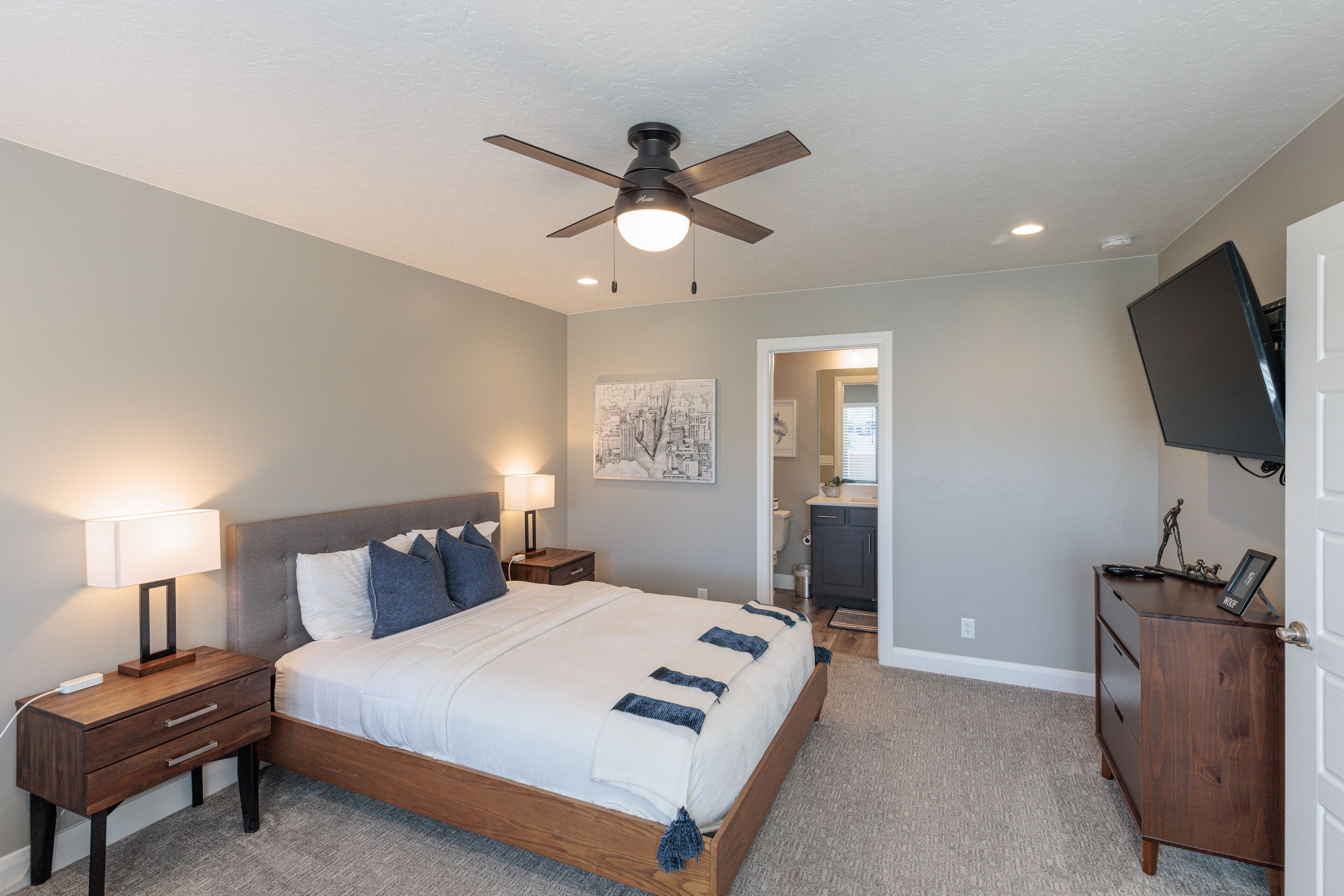 Une chambre à coucher comprenant un lit, des tables de chevet, une commode, un ventilateur de plafond et un téléviseur fixé au mur.