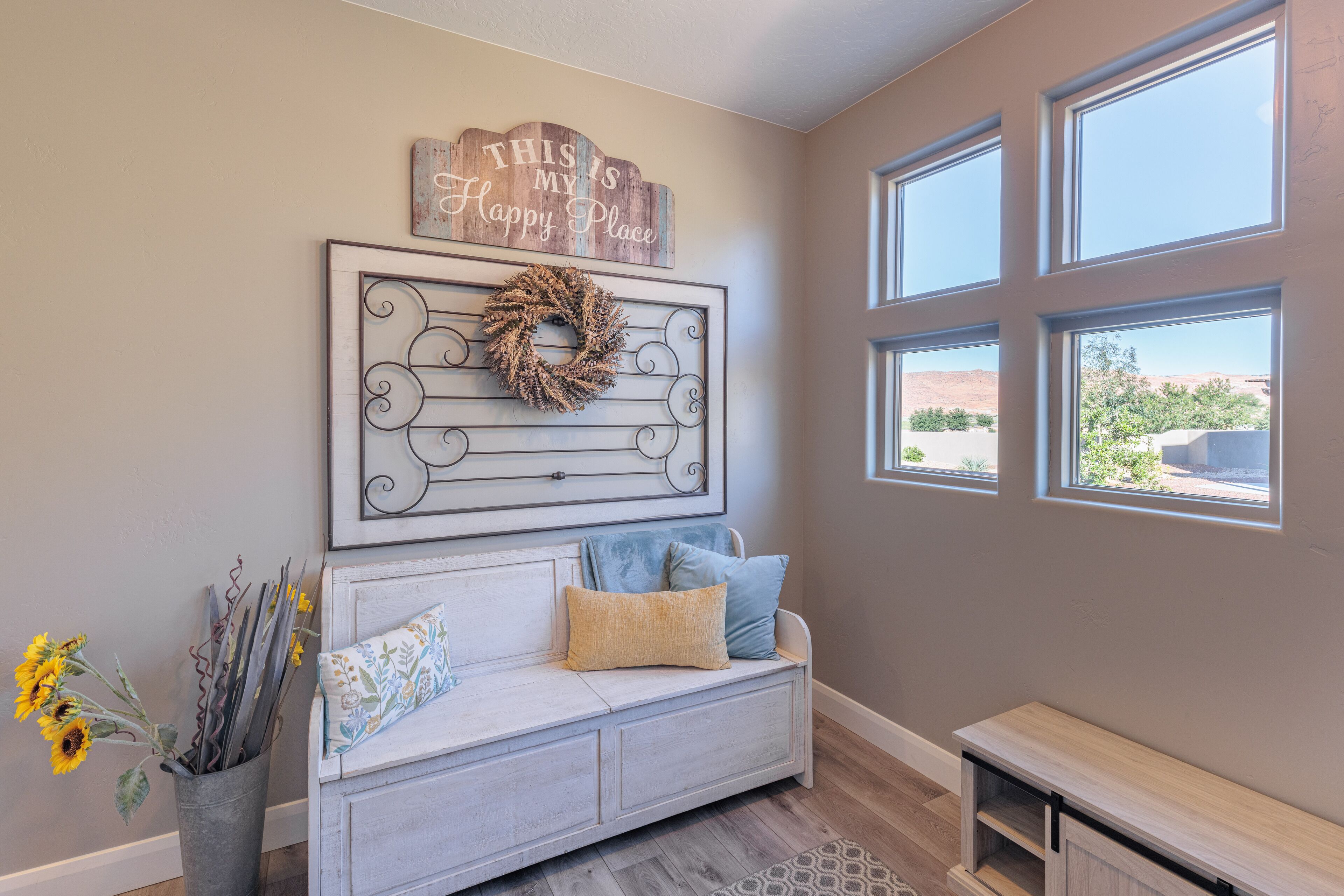 A cozy room with a light gray wall, a wooden sign above a decorative wreath, a white sofa with blue and yellow cushions, and a window with a view of a red rock landscape.